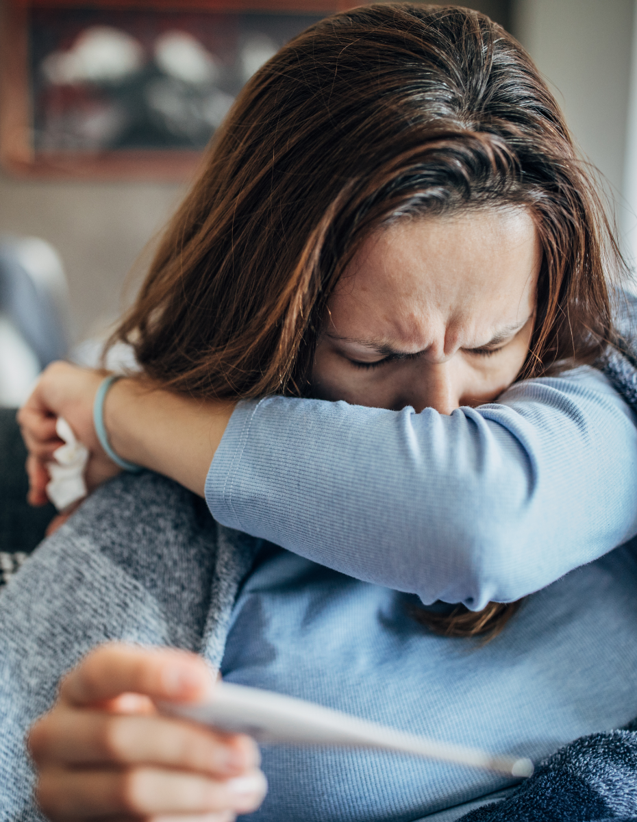 Frau hält Fieberthermometer in der Hand und niest sich in die Armbeuge.